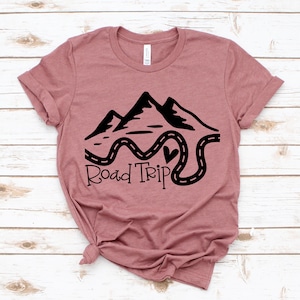 Puede incluir: Una camiseta color malva con un dise&ntilde;o gr&aacute;fico negro de monta&ntilde;as y un camino sinuoso con el texto "Road Trip".