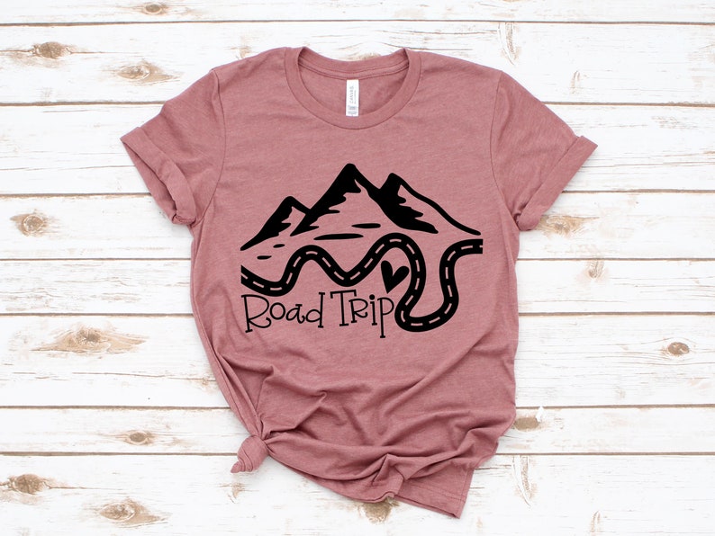 Puede incluir: Una camiseta color malva con un dise&ntilde;o gr&aacute;fico negro de monta&ntilde;as y un camino sinuoso con el texto "Road Trip".