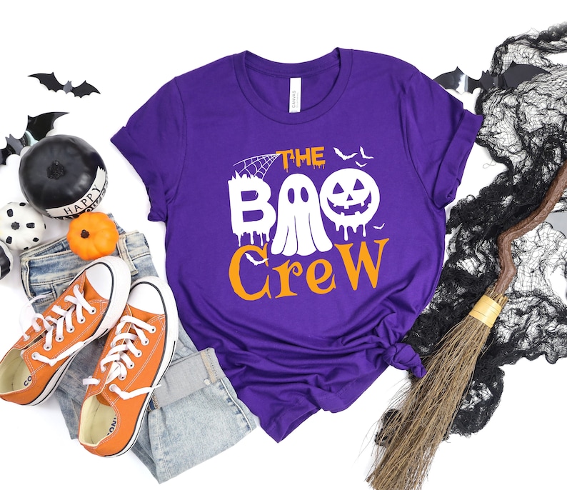 May include: Purple short-sleeved t-shirt with the text "The Boo Crew" in orange and white. A ghost with a dripping face and a jack-o-lantern are part of the design.