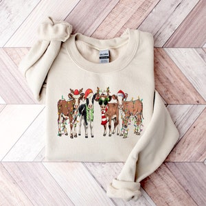 May include: A beige sweatshirt with a graphic of five cows wearing Santa hats and reindeer antlers. The cows are decorated with Christmas lights.