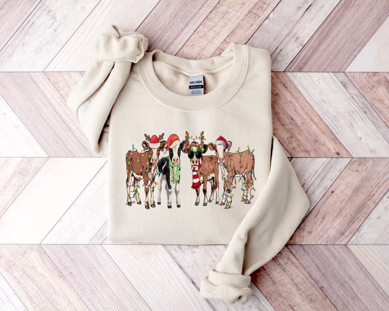May include: A beige sweatshirt with a graphic of five cows wearing Santa hats and reindeer antlers. The cows are decorated with Christmas lights.