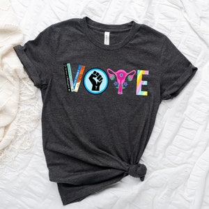 May include: A dark grey t-shirt with the word "VOTE" spelled out in colorful letters. The letters are made up of different images, including a fist, a uterus, and books.