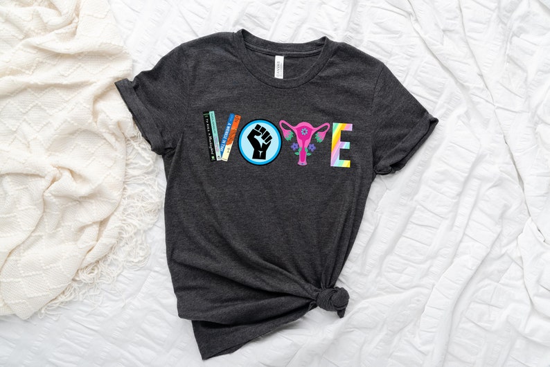 May include: A dark grey t-shirt with the word "VOTE" spelled out in colorful letters. The letters are made up of different images, including a fist, a uterus, and books.