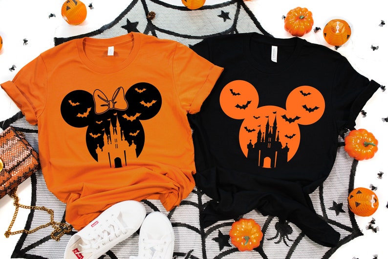May include: Orange and black Halloween-themed t-shirts. One orange shirt features a Minnie Mouse silhouette with a bow, bats, and a castle. The other is black with a Mickey Mouse silhouette, bats, and a castle.