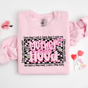 May include: Pink sweatshirt with a black and white checkered design and the text "Motherhood" in pink letters. The text is surrounded by pink flowers and a pink bow. The text reads "Some days I rock it. Some days it rocks me. Either way we're rockin'."