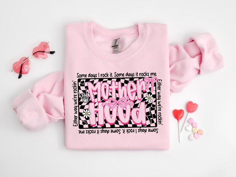 May include: Pink sweatshirt with a black and white checkered design and the text "Motherhood" in pink letters. The text is surrounded by pink flowers and a pink bow. The text reads "Some days I rock it. Some days it rocks me. Either way we're rockin'."