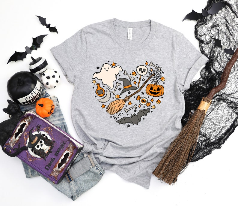 Może przedstawiać: Szary T-shirt z sercowatym wzorem przedstawiającym r&oacute;żne symbole Halloween, w tym ducha, kapelusz czarownicy, miotłę, czaszkę, pajęczynę, dynię, kukurydzę cukrową i słowo "Boo!"