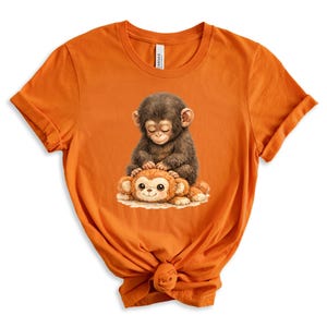 May include: Orange t-shirt featuring a detailed illustration of two cartoon monkeys. One monkey is brown and sitting on top of a smaller, orange monkey. The design is centered on the front of the shirt, which is made of a soft material.