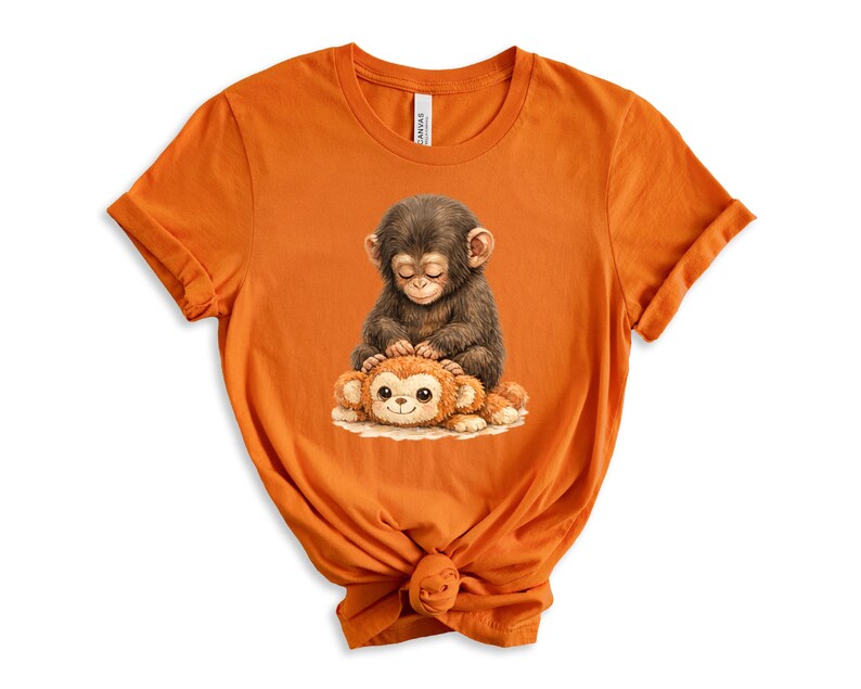 May include: Orange t-shirt featuring a detailed illustration of two cartoon monkeys. One monkey is brown and sitting on top of a smaller, orange monkey. The design is centered on the front of the shirt, which is made of a soft material.