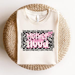 May include: A white t-shirt with a black and white checkered design featuring the text "Motherhood" in pink with a pink bow. The text around the design reads "Some days I rock it. Some days it rocks me. Either way we're rockin'."