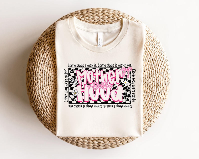 May include: A white t-shirt with a black and white checkered design featuring the text "Motherhood" in pink with a pink bow. The text around the design reads "Some days I rock it. Some days it rocks me. Either way we're rockin'."