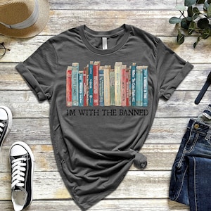 K&ouml;nnte beinhalten: Ein dunkelgraues T-Shirt mit einem Aufdruck eines B&uuml;cherregals voller verbotener B&uuml;cher. Der Text "I'm with the banned" ist unter dem B&uuml;cherregal-Motiv gedruckt.