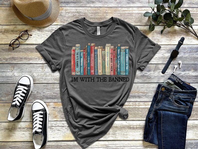 K&ouml;nnte beinhalten: Ein dunkelgraues T-Shirt mit einem Aufdruck eines B&uuml;cherregals voller verbotener B&uuml;cher. Der Text "I'm with the banned" ist unter dem B&uuml;cherregal-Motiv gedruckt.