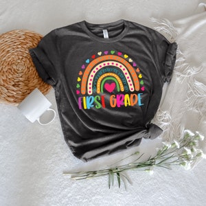 Peut inclure: Un t-shirt gris fonc&eacute; avec un motif arc-en-ciel et le texte "FIRST GRADE" en lettres color&eacute;es. L'arc-en-ciel a un c&oelig;ur rose au centre et est entour&eacute; de petits c&oelig;urs.