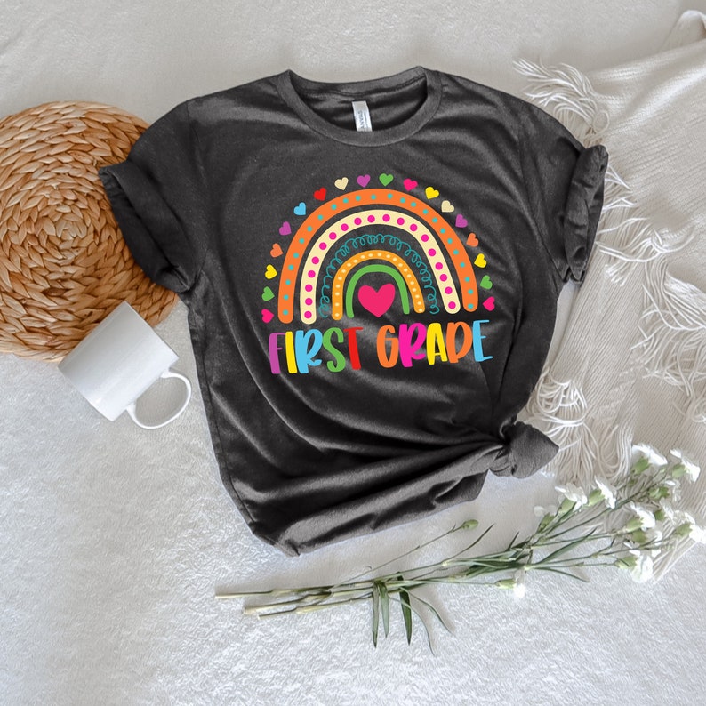 Peut inclure: Un t-shirt gris fonc&eacute; avec un motif arc-en-ciel et le texte "FIRST GRADE" en lettres color&eacute;es. L'arc-en-ciel a un c&oelig;ur rose au centre et est entour&eacute; de petits c&oelig;urs.