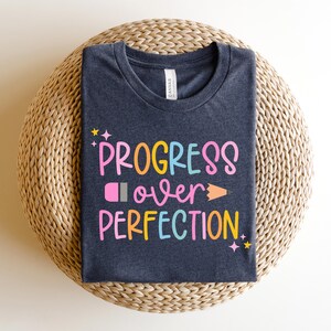 May include: Dark navy blue t-shirt with the words "PROGRESS over PERFECTION" in colorful, playful lettering. The design includes a pencil and stars. The shirt is displayed on a woven, circular surface.