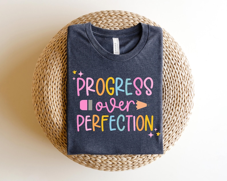 May include: Dark navy blue t-shirt with the words "PROGRESS over PERFECTION" in colorful, playful lettering. The design includes a pencil and stars. The shirt is displayed on a woven, circular surface.