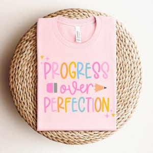 May include: A light pink t-shirt with the words "PROGRESS over PERFECTION" in colorful letters and a pencil graphic. The shirt is displayed on a woven, circular surface.