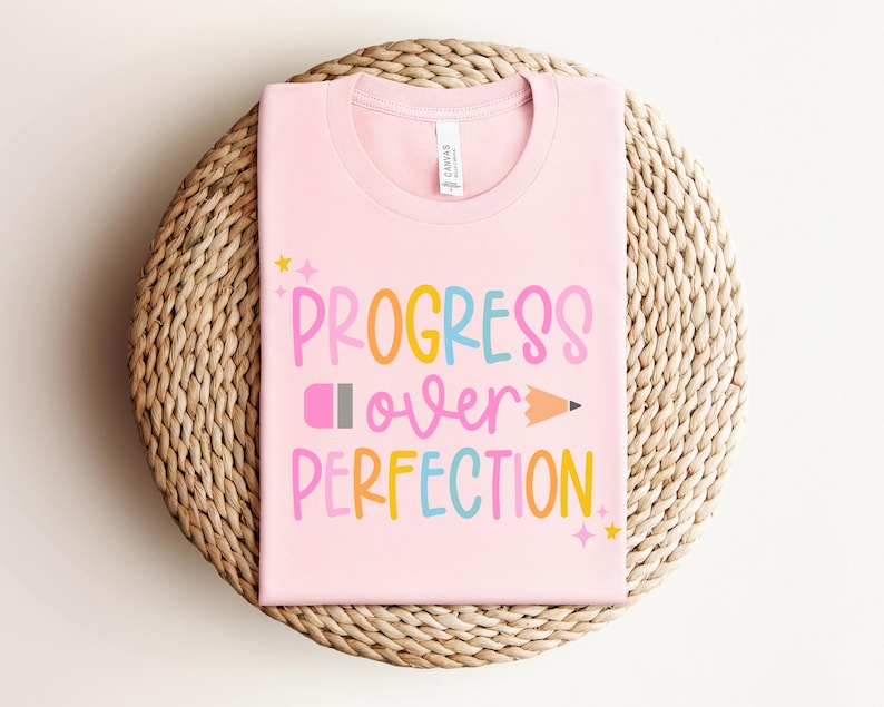 May include: A light pink t-shirt with the words "PROGRESS over PERFECTION" in colorful letters and a pencil graphic. The shirt is displayed on a woven, circular surface.