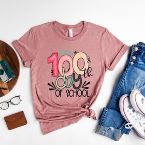 May include: A pink t-shirt with the text "100th day of school" in colorful, stylized lettering.