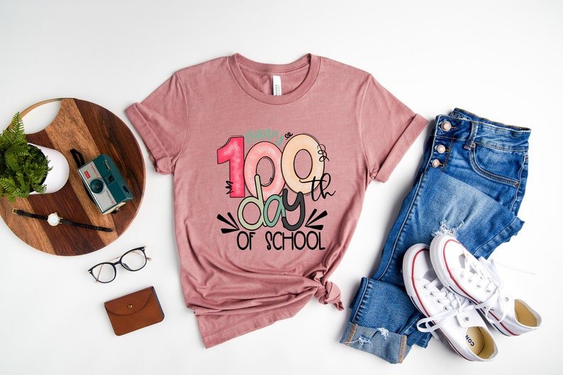 May include: A pink t-shirt with the text "100th day of school" in colorful, stylized lettering.