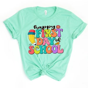 May include: A light green t-shirt with the text "happy first day of school" in colorful, retro-style lettering. A cartoon pencil with a red eraser is next to the text.
