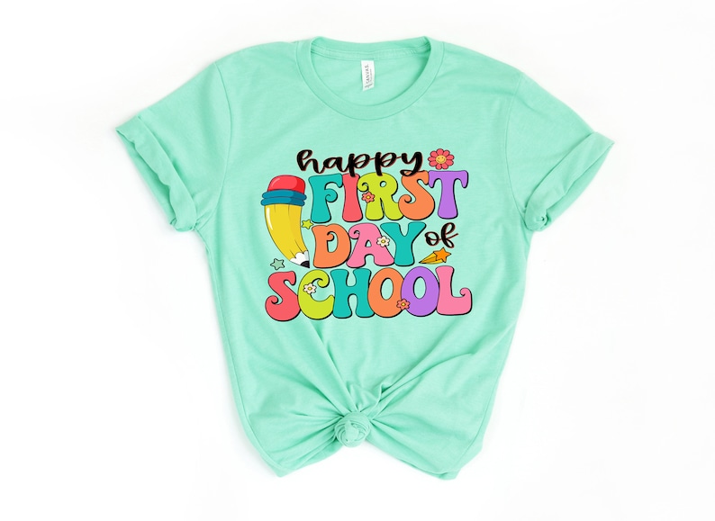 May include: A light green t-shirt with the text "happy first day of school" in colorful, retro-style lettering. A cartoon pencil with a red eraser is next to the text.