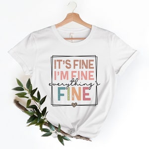 May include: White t-shirt with a graphic print that says "It's fine I'm fine everything's fine" in a colorful retro font with a small heart.