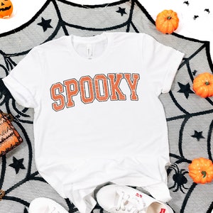 May include: White t-shirt with the word "SPOOKY" printed in orange on the front. The shirt is surrounded by a black spiderweb, orange pumpkins, and black spiders.