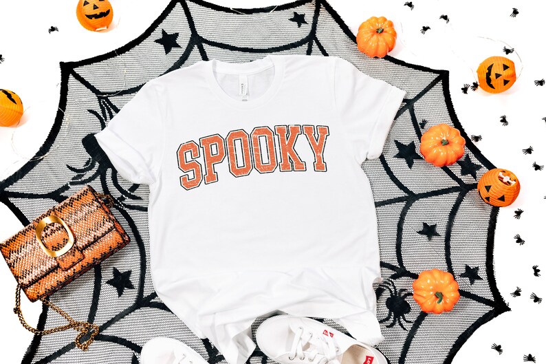 May include: White t-shirt with the word "SPOOKY" printed in orange on the front. The shirt is surrounded by a black spiderweb, orange pumpkins, and black spiders.