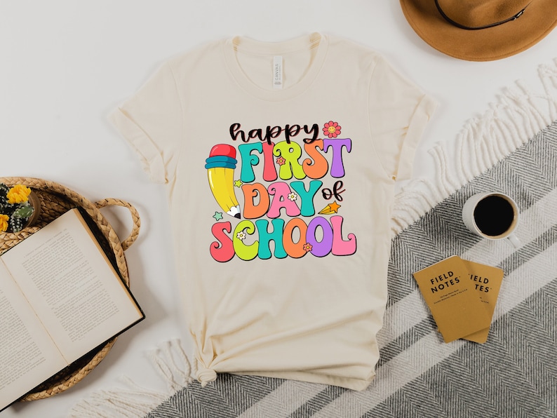 May include: A beige t-shirt with the text "happy first day of school" in colorful, retro-style lettering. A cartoon pencil is featured next to the text.