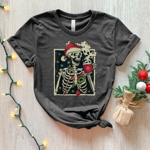 May include: A dark gray t-shirt with a graphic of a skeleton wearing a Santa hat and holding a mug of hot cocoa. The skeleton is surrounded by Christmas decorations, including candy canes, holly, and snowflakes.