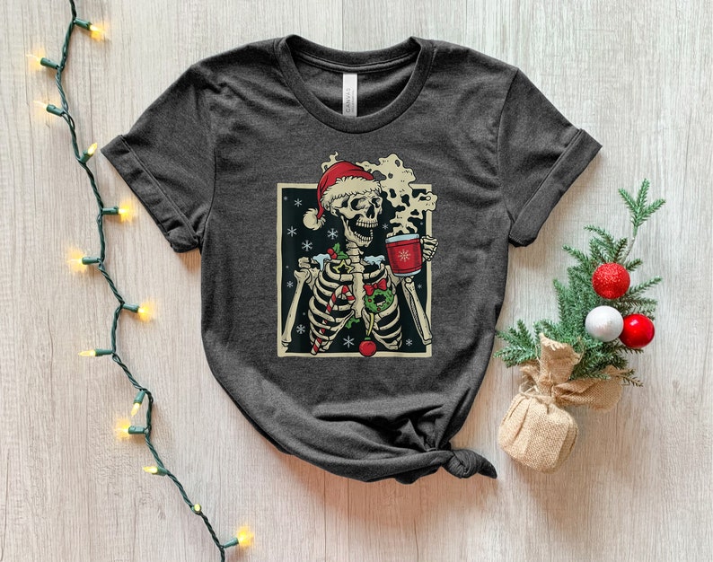 May include: A dark gray t-shirt with a graphic of a skeleton wearing a Santa hat and holding a mug of hot cocoa. The skeleton is surrounded by Christmas decorations, including candy canes, holly, and snowflakes.