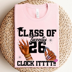 May include: A light pink t-shirt with the text "CLASS OF Twenty 26 CLOCK ITTTT!!" in black. The design includes illustrations of hands with long, manicured nails, a graduation cap, and a kiss mark.