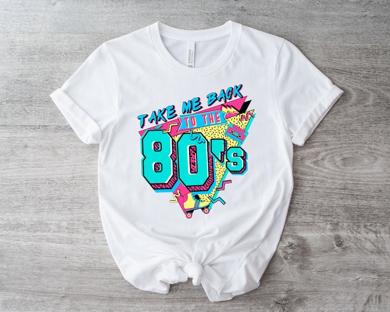 Op de afbeelding: Wit T-shirt met een grafisch ontwerp dat "Take Me Back to the 80's" zegt in een kleurrijke, retro stijl. Het ontwerp bevat een cassettebandje, rol schaatsen en geometrische vormen.