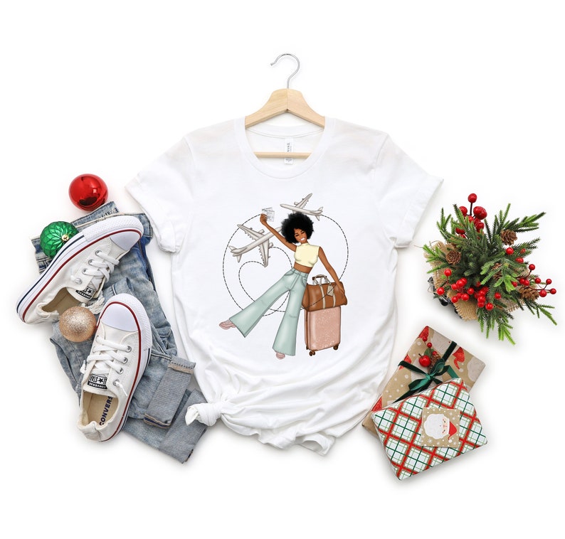 Puede incluir: Camiseta blanca con un gr&aacute;fico de una mujer con cabello casta&ntilde;o y una camisa amarilla sosteniendo una maleta y saludando a los aviones.