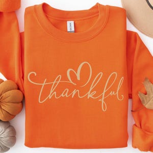 May include: A bright orange long-sleeve crewneck sweatshirt featuring the word thankful written in a light tan cursive script with a heart shape integrated into the lettering. The garment has a ribbed collar and a standard fit design.