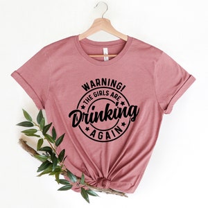 May include: A dusty rose t-shirt with rolled sleeves, hanging on a wooden hanger. The shirt features a black circular graphic with the words "WARNING! THE GIRLS ARE Drinking AGAIN" in a bold, playful font.