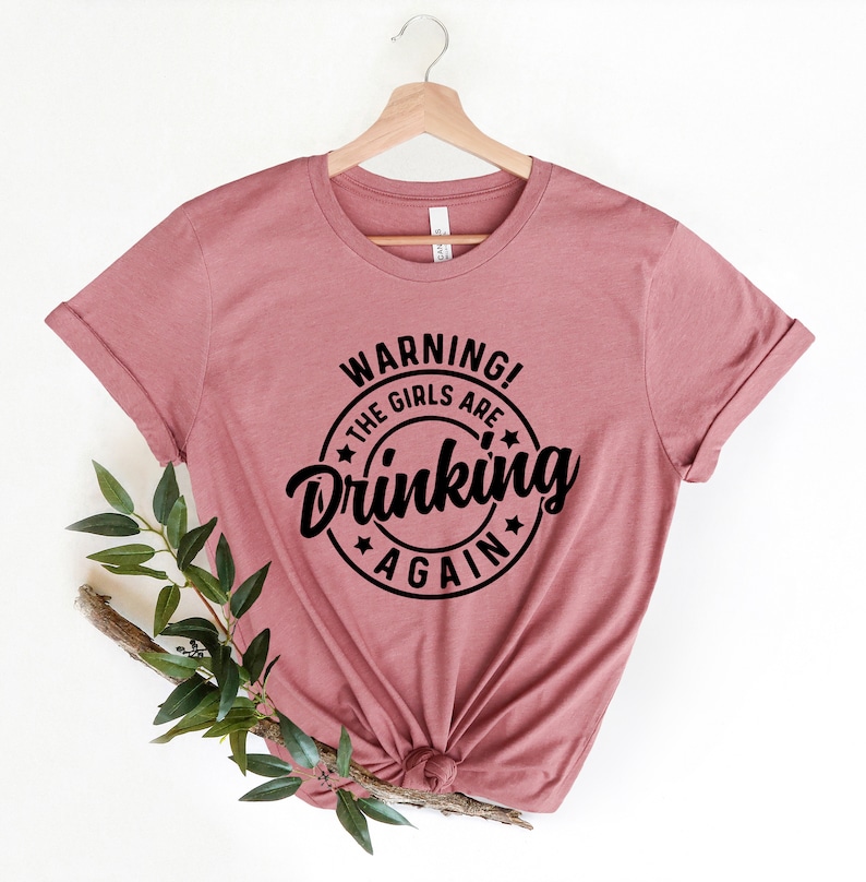 May include: A dusty rose t-shirt with rolled sleeves, hanging on a wooden hanger. The shirt features a black circular graphic with the words "WARNING! THE GIRLS ARE Drinking AGAIN" in a bold, playful font.