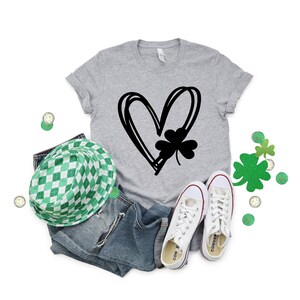 May include: A gray t-shirt with a black graphic of a heart with a four-leaf clover inside. The shirt is paired with blue jeans, a green and white checkered hat, and white sneakers.