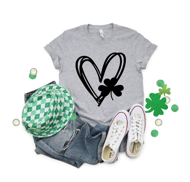 May include: A gray t-shirt with a black graphic of a heart with a four-leaf clover inside. The shirt is paired with blue jeans, a green and white checkered hat, and white sneakers.