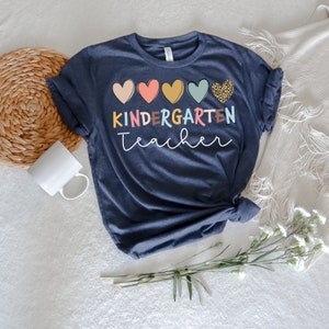 K&ouml;nnte beinhalten: Ein marineblaues T-Shirt mit dem Text "KINDERGARTEN teacher" in bunten Buchstaben. Das Shirt zeigt eine Reihe von f&uuml;nf bunten Herzen, darunter ein Herz mit Leopardenmuster.