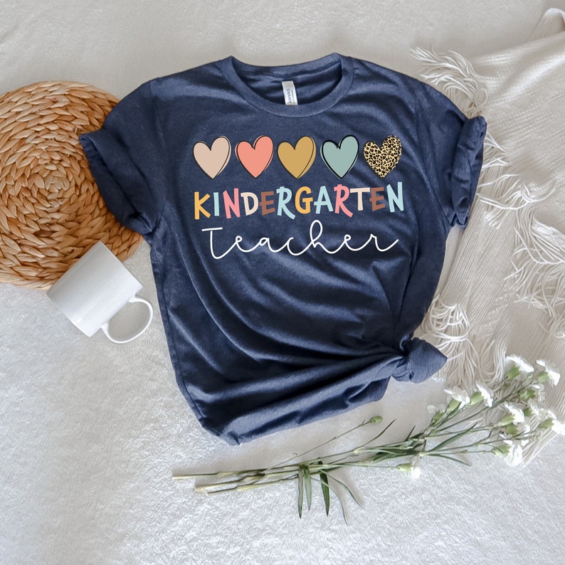 K&ouml;nnte beinhalten: Ein marineblaues T-Shirt mit dem Text "KINDERGARTEN teacher" in bunten Buchstaben. Das Shirt zeigt eine Reihe von f&uuml;nf bunten Herzen, darunter ein Herz mit Leopardenmuster.