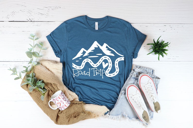 Puede incluir: Una camiseta azul turquesa con un gr&aacute;fico blanco de monta&ntilde;as y un camino sinuoso con un coraz&oacute;n en el medio. El texto "Road Trip" est&aacute; escrito debajo del gr&aacute;fico.