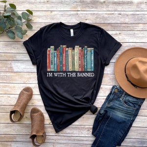K&ouml;nnte beinhalten: Schwarzes T-Shirt mit einem Grafikdruck eines B&uuml;cherregals voller verbotener B&uuml;cher. Der Text "I'm with the banned" ist unter dem B&uuml;cherregal-Grafikdruck gedruckt.