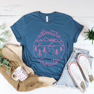 May include: A teal blue t-shirt with a pink graphic design of mountains, a river, and a sun. The graphic design is in a circle with the text "adventure awaits" written in a cursive font.