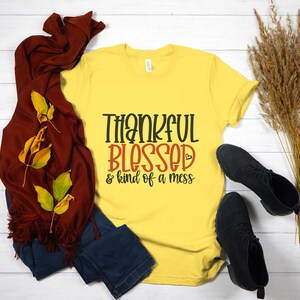 May include: Yellow t-shirt with the text "Thankful Blessed & kind of a mess" in black and red lettering. The shirt is on a white wooden surface with a brown scarf, blue jeans, and dried grass.