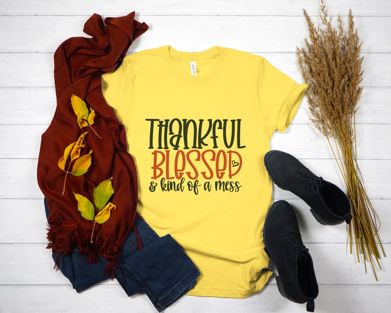 May include: Yellow t-shirt with the text "Thankful Blessed & kind of a mess" in black and red lettering. The shirt is on a white wooden surface with a brown scarf, blue jeans, and dried grass.