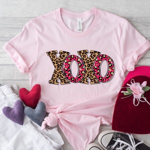May include: Pink t-shirt with a graphic of the letters XOXO in a leopard print design. The letters are outlined in black and have a pink and gold interior.