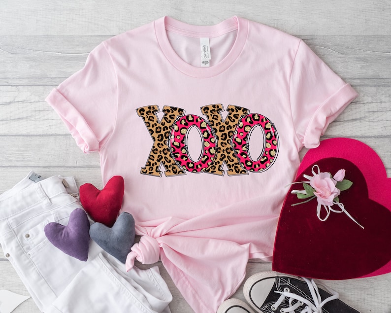 May include: Pink t-shirt with a graphic of the letters XOXO in a leopard print design. The letters are outlined in black and have a pink and gold interior.